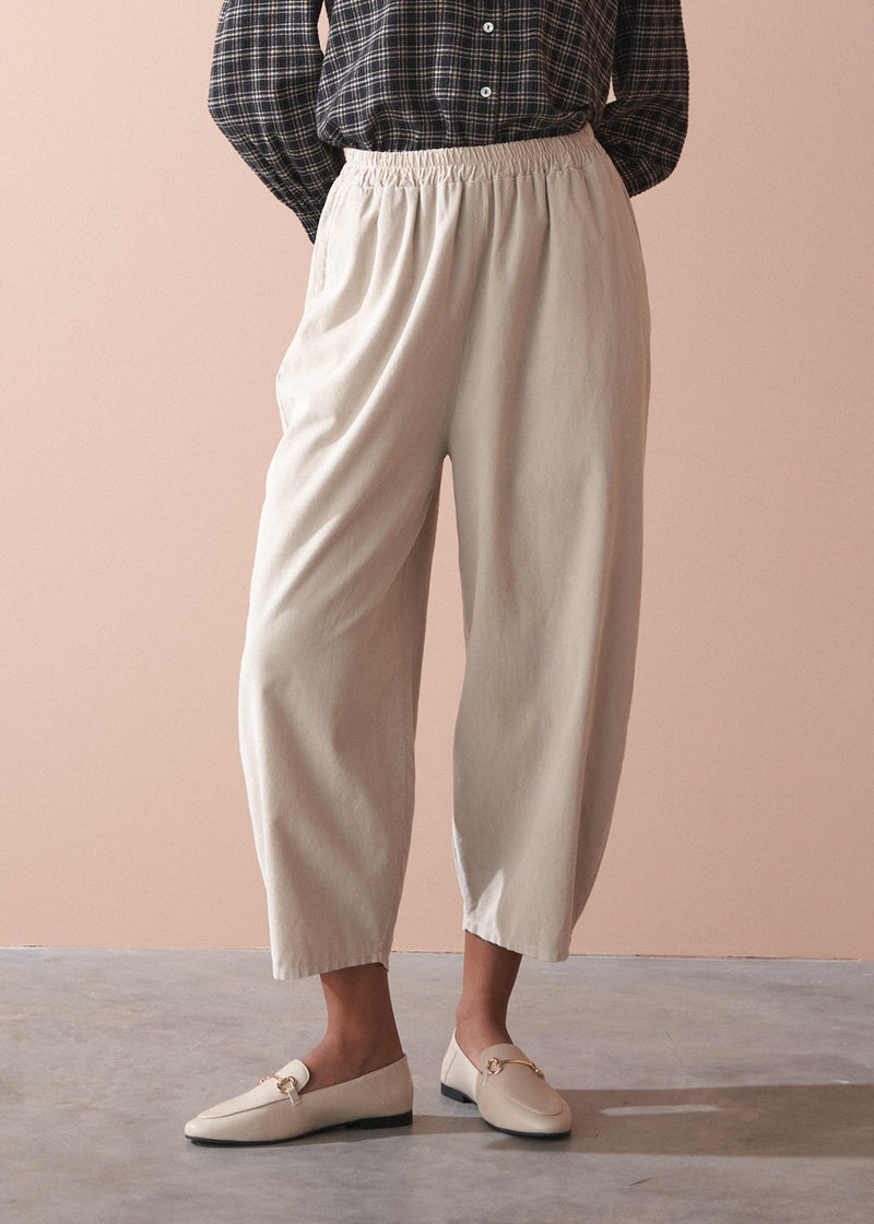 Woman wearing a check shirt and beige corduroy barrel leg trousers against a beige background