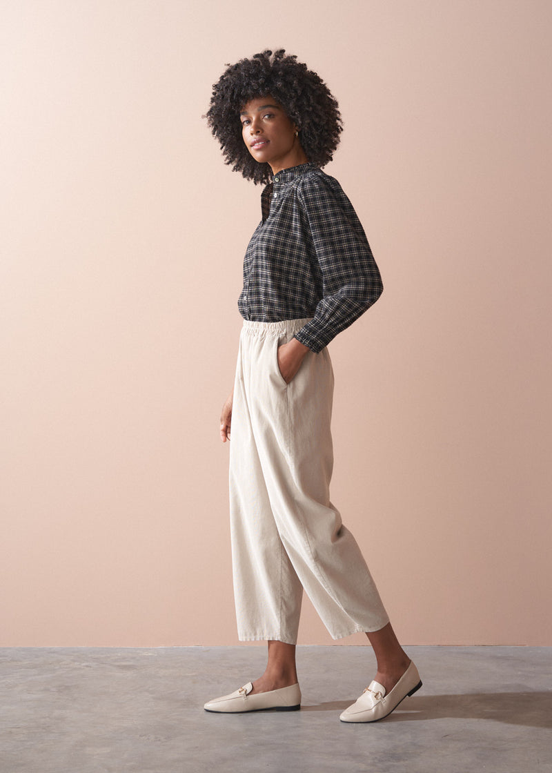 Woman wearing a check shirt and beige corduroy barrel leg trousers against a beige background