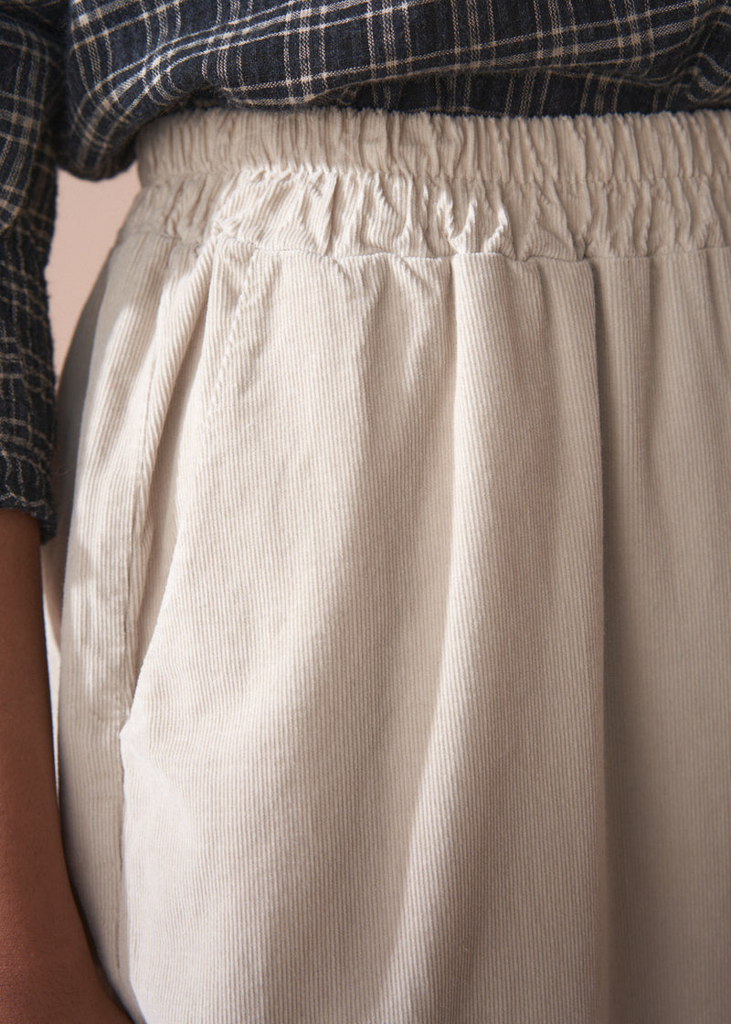 Woman wearing a check shirt and beige corduroy barrel leg trousers against a beige background
