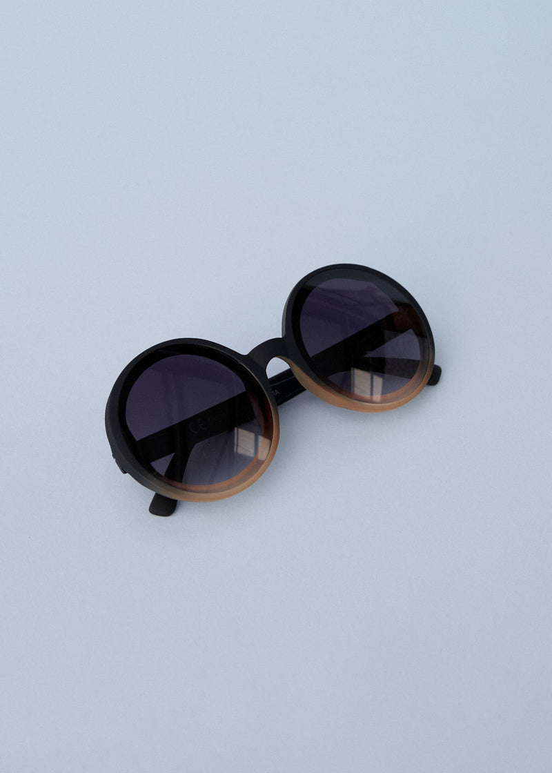 A pair of chunky circular sunglasses with black and pink frames and blue lenses