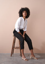 Woman sitting on a wooden stool wearing a white blouse and black trousers against a beige background