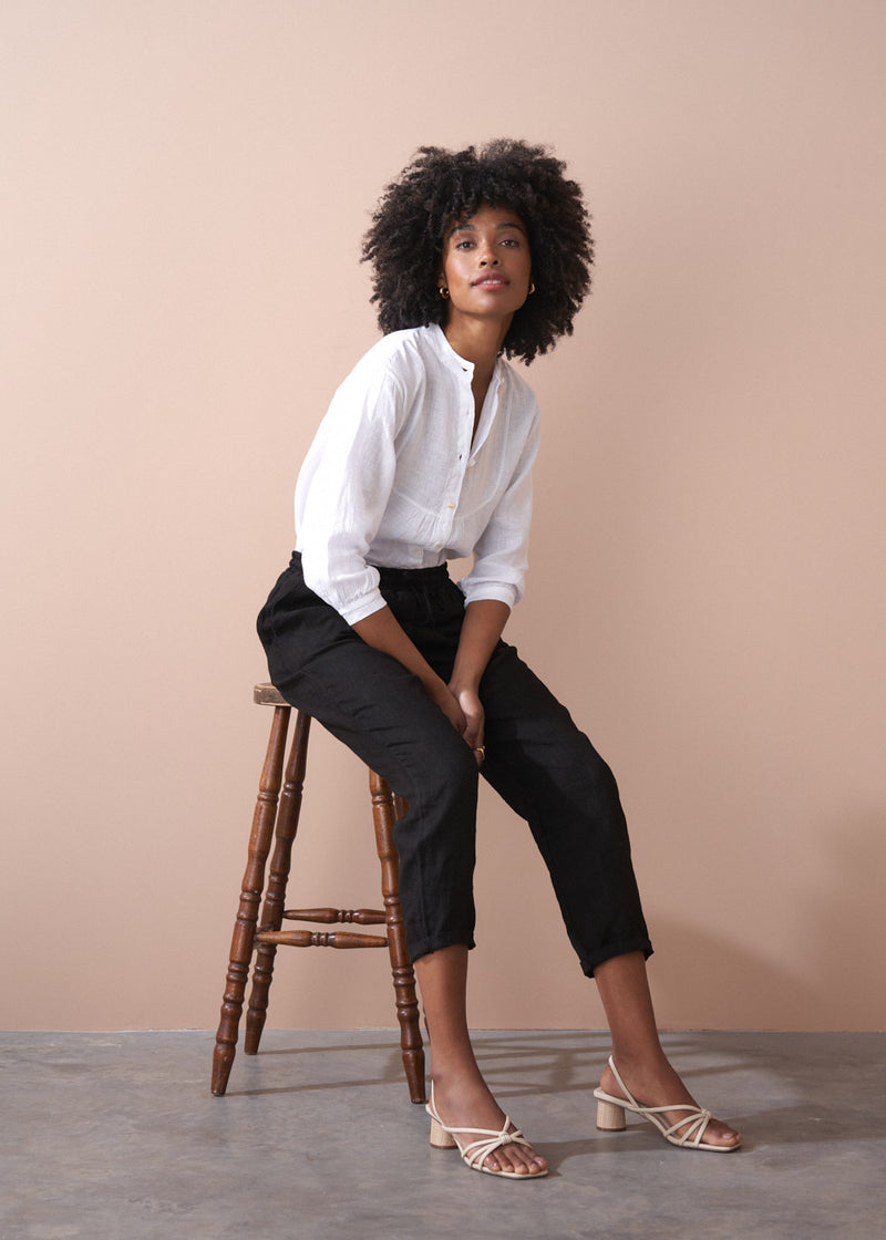 Woman sitting on a wooden stool wearing a white blouse and black trousers against a beige background
