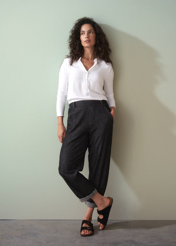 Woman wearing a white shirt and dark jeans against a plain wall.