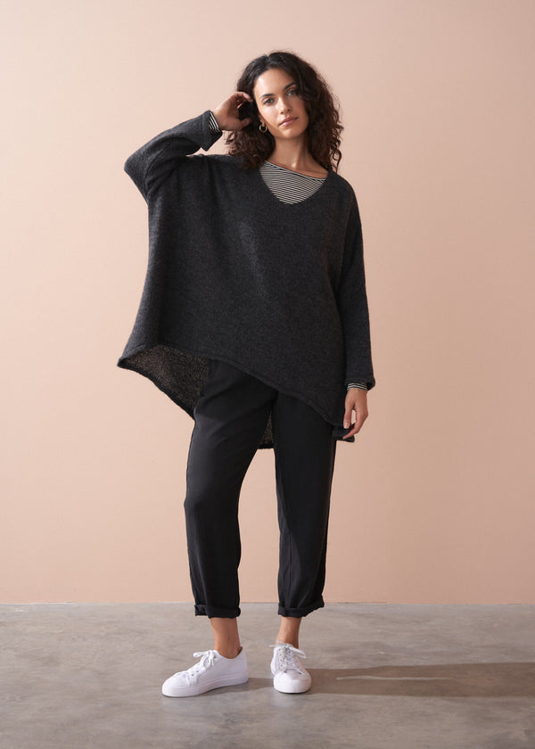 A woman wearing a dark grey oversized sweater over a striped top and black trousers with white trainers on a beige background