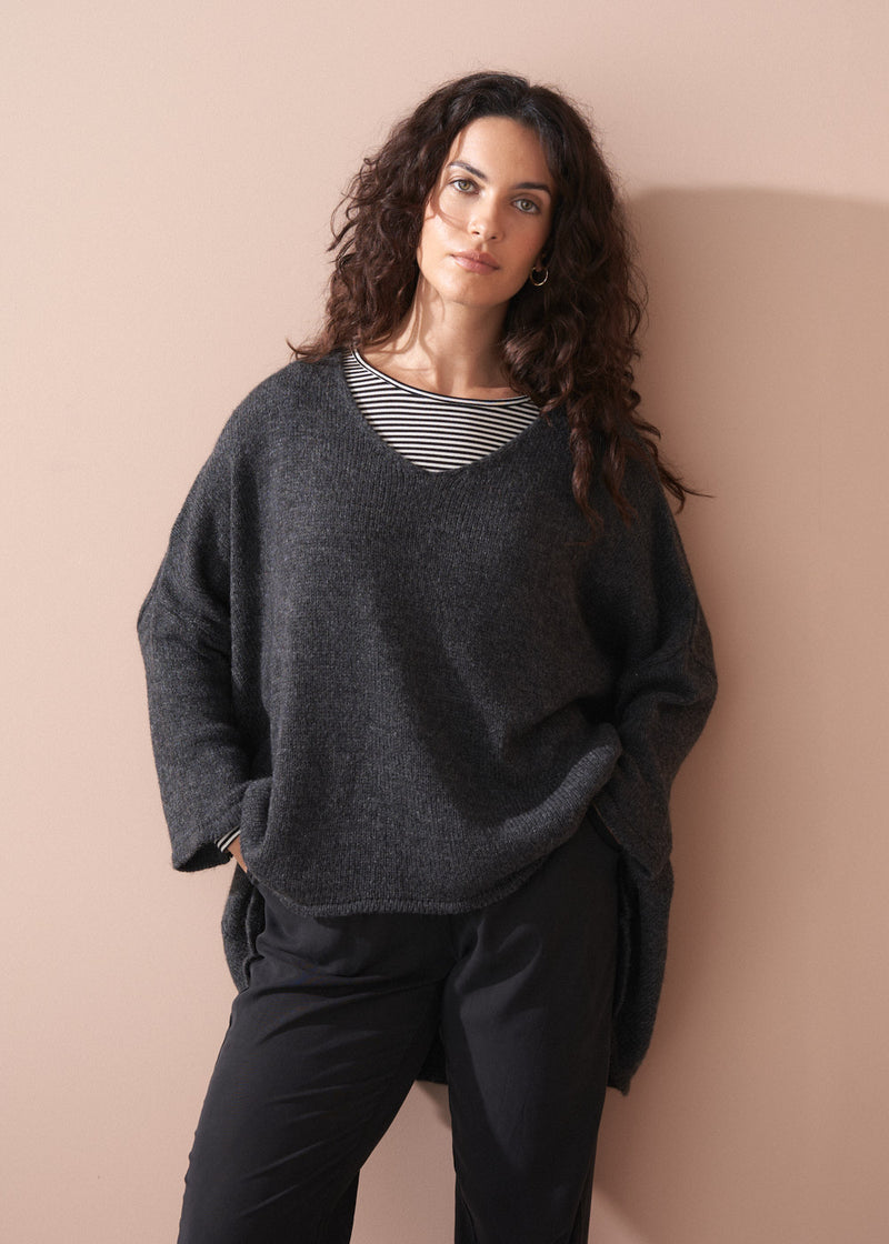 A woman wearing a dark grey oversized sweater over a striped top and black trousers with white trainers on a beige background