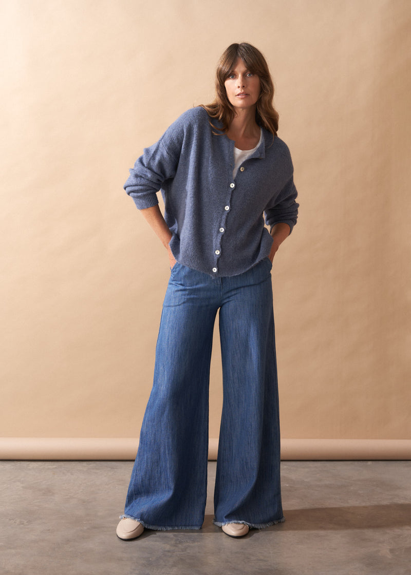 Model wearing light blue wool blend cardigan with mother of pearl buttons and wide leg jeans against a plain background