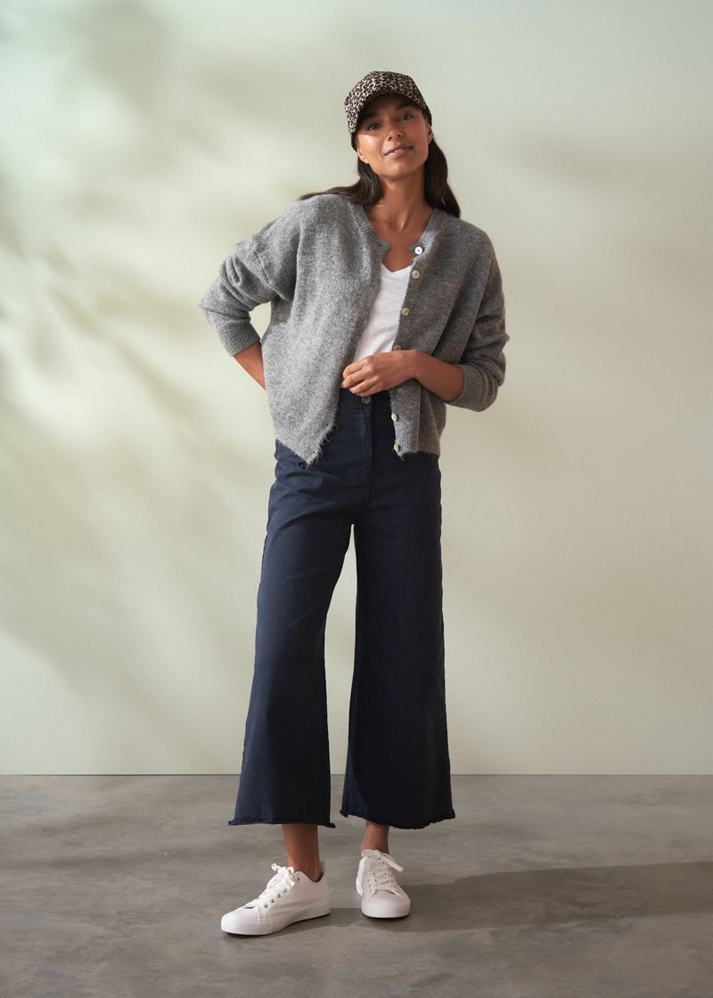 Woman wearing a grey wool cardigan over a white shirt and dark trousers on a plain background
