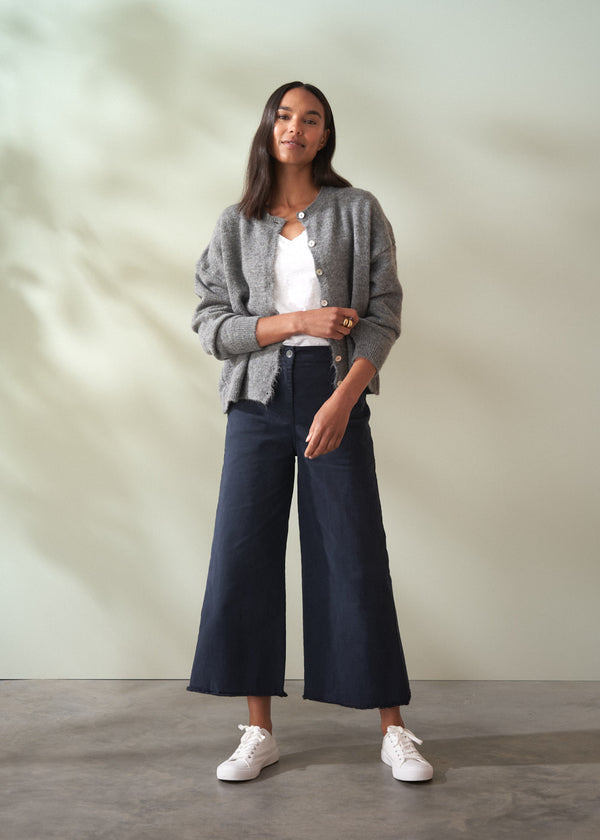 Woman wearing a grey wool cardigan over a white shirt and dark trousers on a plain background