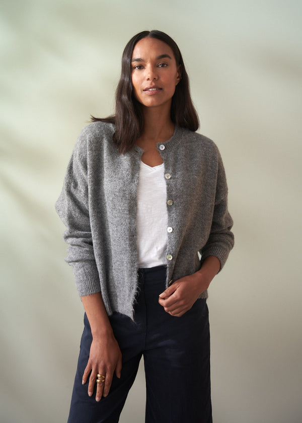 Woman wearing a grey wool cardigan over a white shirt and dark trousers on a plain background