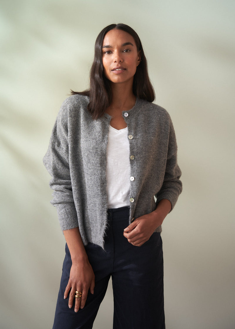 Woman wearing a grey wool cardigan over a white shirt and dark trousers on a plain background