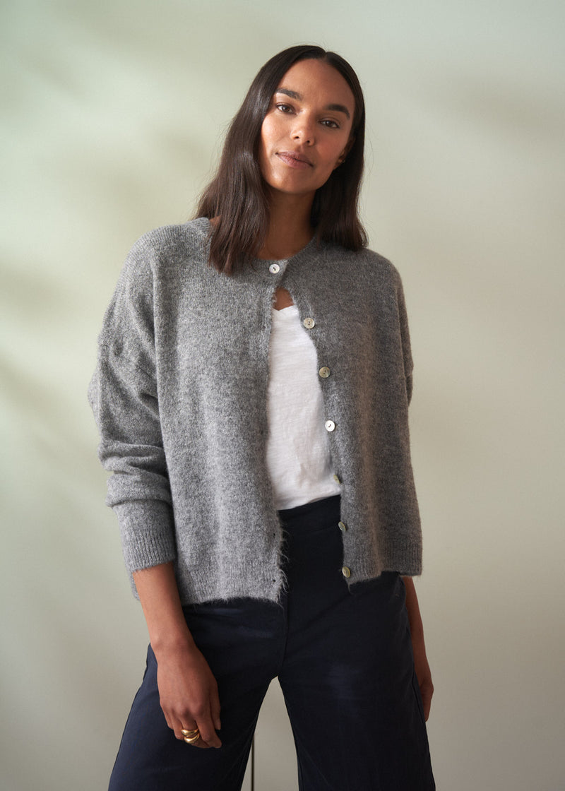 Woman wearing a grey wool cardigan over a white shirt and dark trousers on a plain background