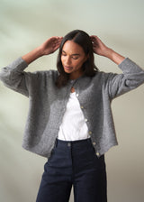 Woman wearing a grey wool cardigan over a white shirt and dark trousers on a plain background