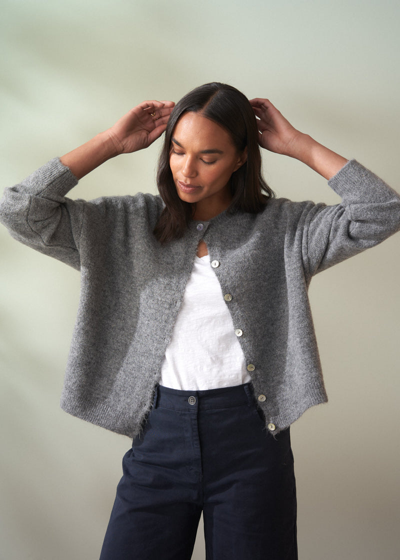 Woman wearing a grey wool cardigan over a white shirt and dark trousers on a plain background