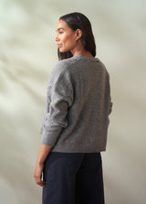 Woman wearing a grey wool cardigan over a white shirt and dark trousers on a plain background