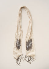 An off-white scarf with fringe and silver beaded geometric patterns, laid flat on a light beige background.
