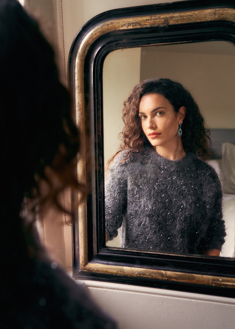 Woman wearing a dark grey textured sweater with sequins and statement earrings, looking into an antique mirror