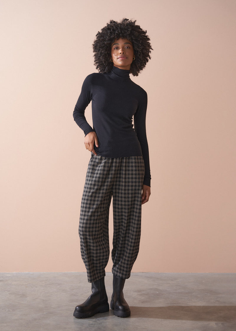 Person wearing a black turtleneck and checkered barrel leg trousers against a beige background