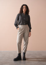 Woman wearing a black and white checkered shirt, beige slim fit trousers, and black boots against a beige background