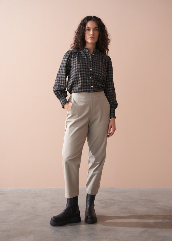 Woman wearing a black and white checkered shirt, beige slim fit trousers, and black boots against a beige background