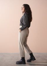 Woman wearing a black and white checkered shirt, beige slim fit trousers, and black boots against a beige background