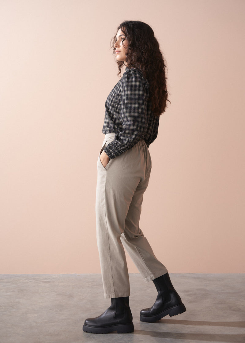 Woman wearing a black and white checkered shirt, beige slim fit trousers, and black boots against a beige background