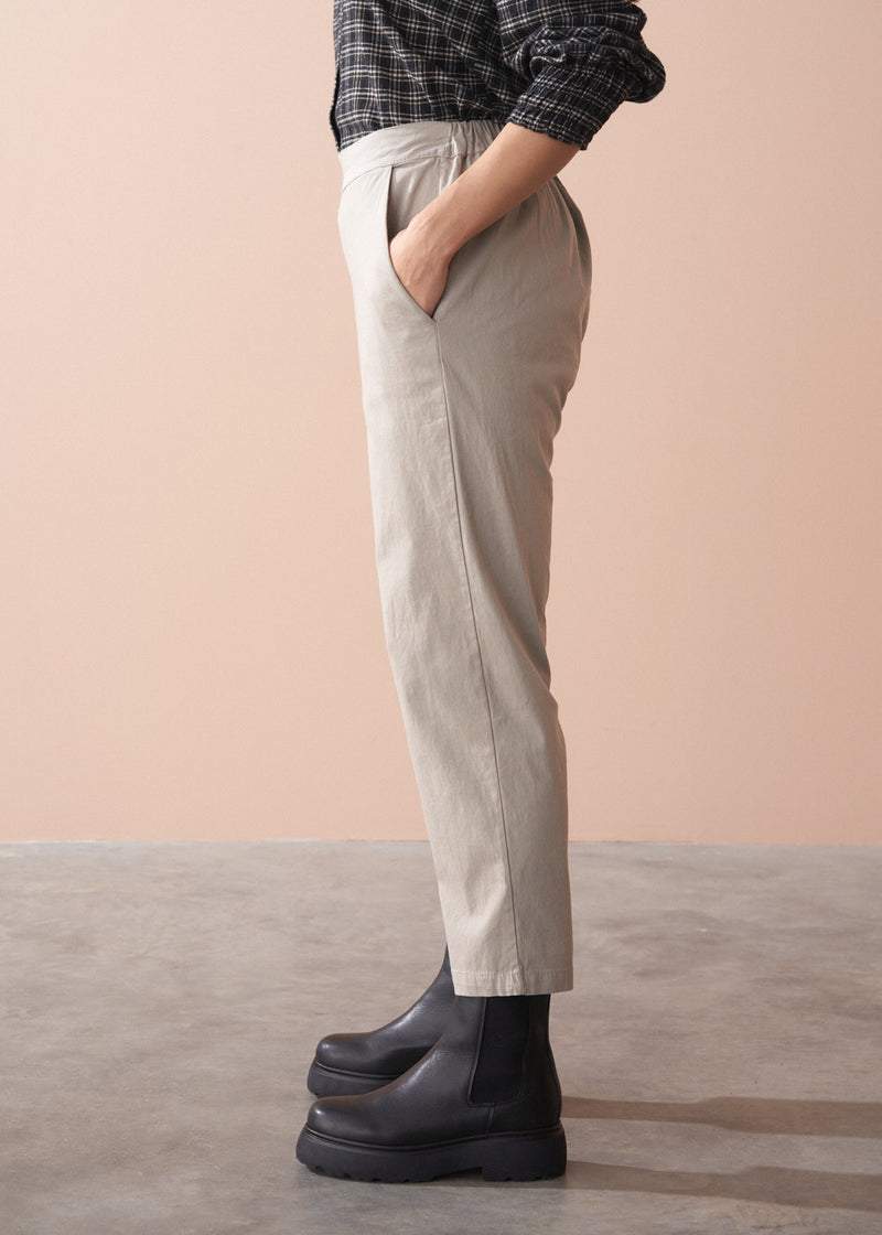 Woman wearing a black and white checkered shirt, beige slim fit trousers, and black boots against a beige background