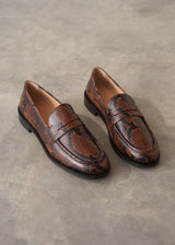 A pair of brown and black snakeskin-patterned penny loafers on a textured gray concrete surface.