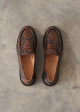 A pair of women's penny loafers with a brown and black snakeskin pattern, shown from a top-down view on a textured gray concrete surface.