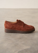 A single brown suede oxford-style shoe with a low platform sole, shown in profile against a neutral gray concrete floor and light beige wall background.