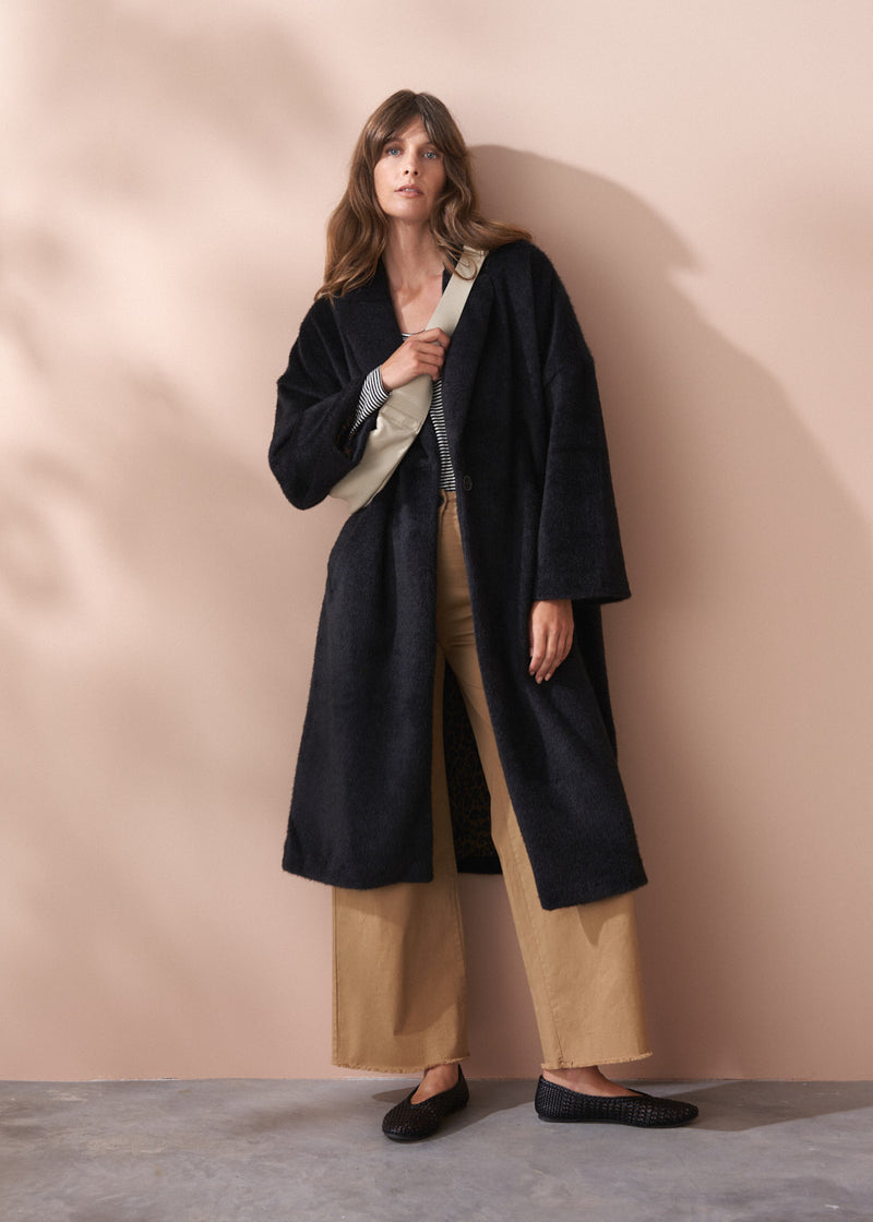 Woman wearing a black fur coat and neutral wide leg trousers against a beige background