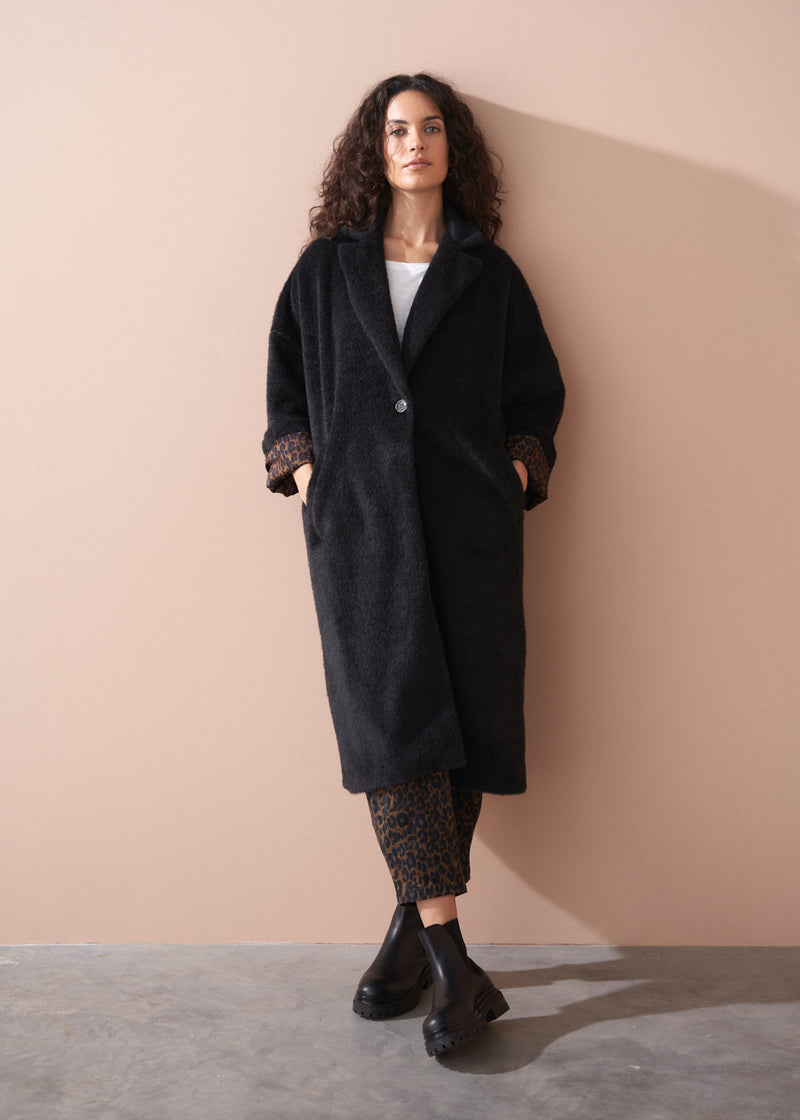Woman wearing a long black fur coat, white top, and leopard print trousers against a beige wall.