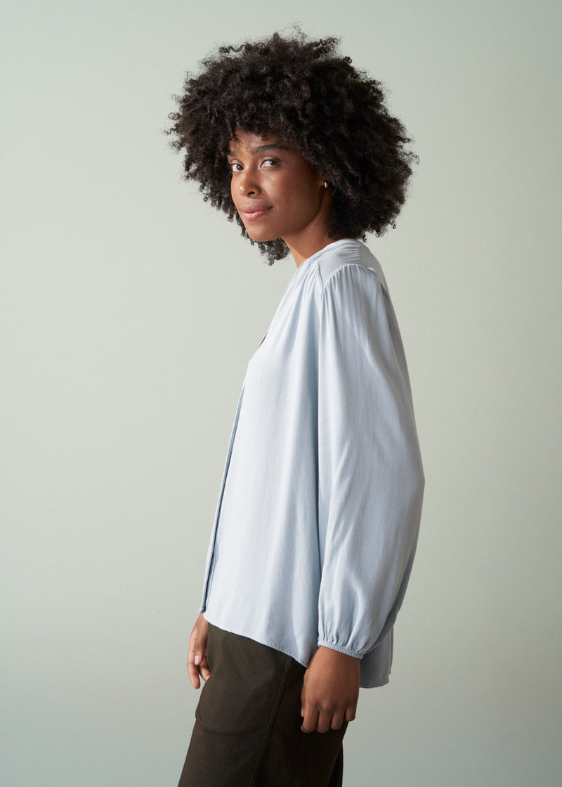 Woman wearing a light blue shirt and dark green trousers with a plain background