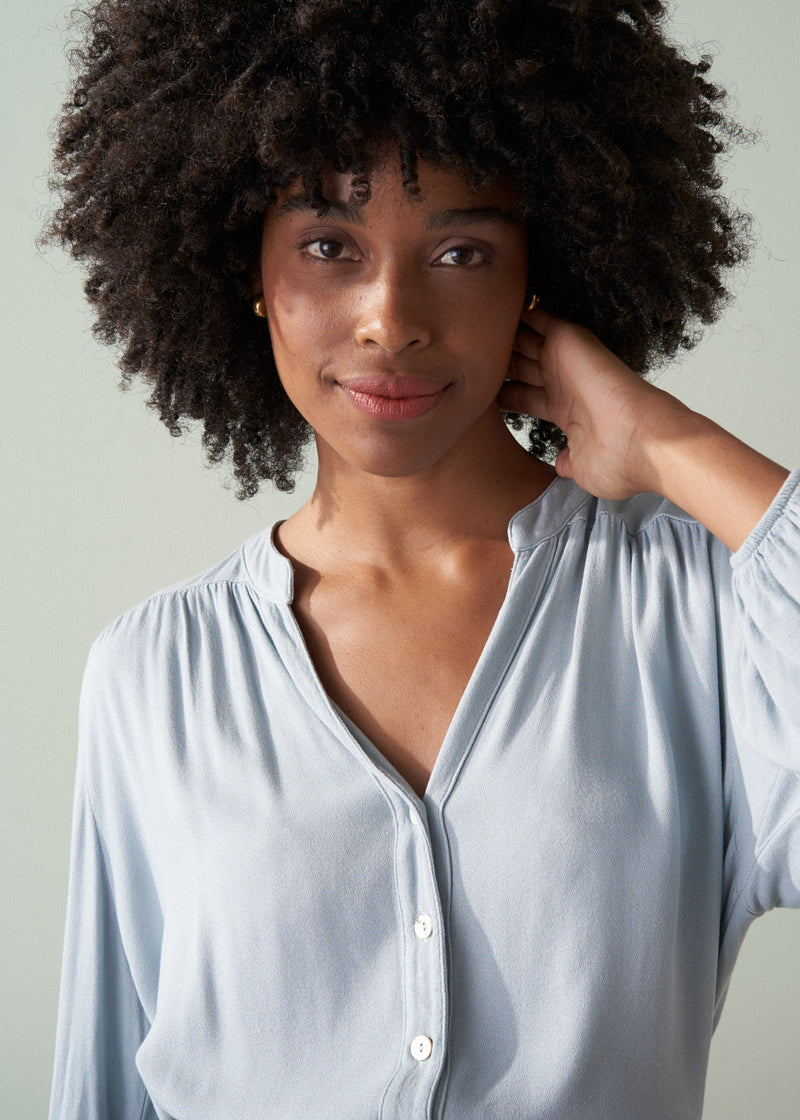 Woman wearing a light blue shirt and dark green trousers with a plain background