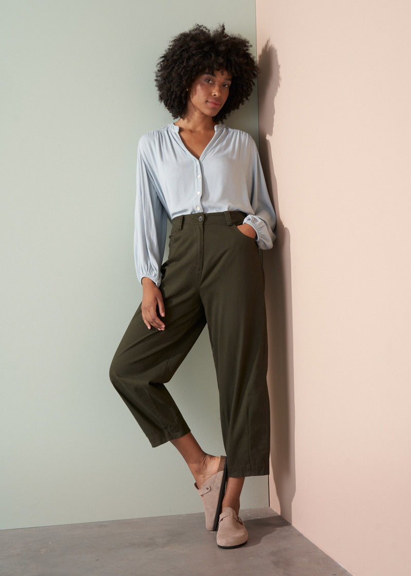 Woman wearing a light blue shirt and dark green trousers with a plain background