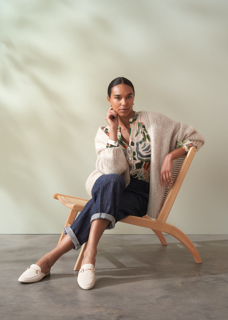 Woman sitting on a wooden chair wearing a patterned top, cardigan, and pants.