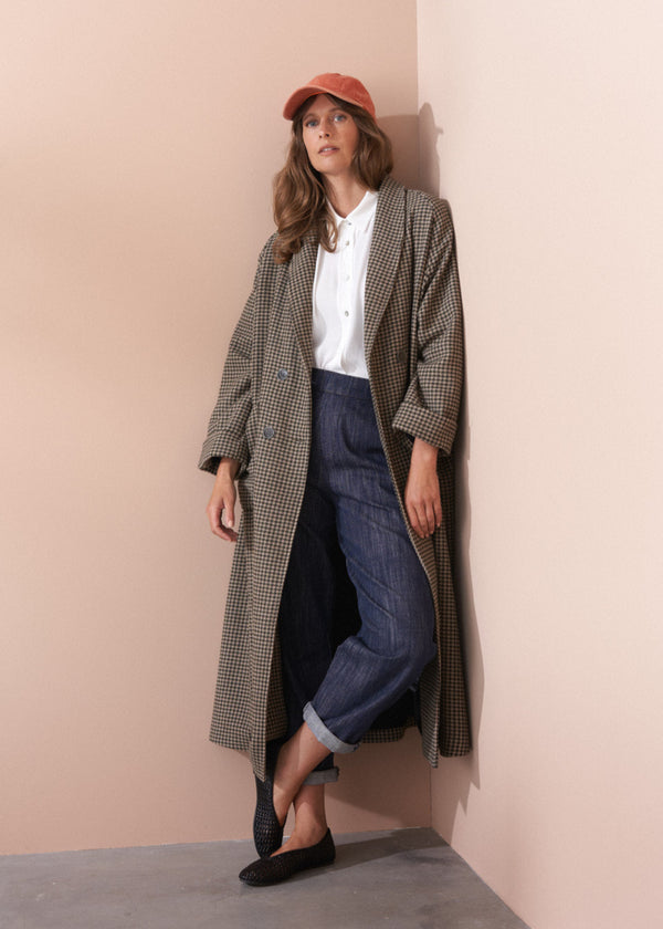 Woman wearing a long check coat, white shirt, and jeans against a beige wall.