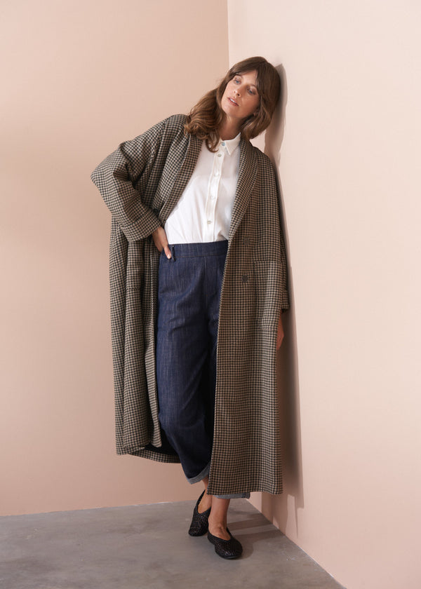 Woman wearing a long checkered coat against a plain wall.