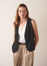 Woman wearing a black vest over a white top and gold satin trousers on a plain background