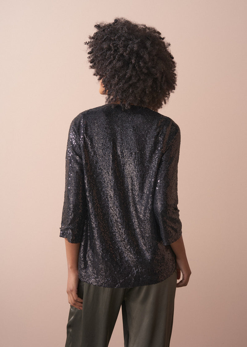 Woman wearing a black sequin jacket, black satin camisole and green satin trousers against a beige background