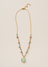 A delicate gold chain necklace featuring a rectangular light green pendant and small multicolored beads with tiny gold charms, displayed flat against a textured off-white background.