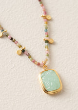 A gold-tone beaded necklace featuring a light green jadeite pendant with an embossed floral pattern, displayed on a textured off-white background.