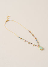 A delicate gold chain necklace featuring a small beaded strand with tiny multicolored stones and a polished light green pendant, displayed on a warm beige background.