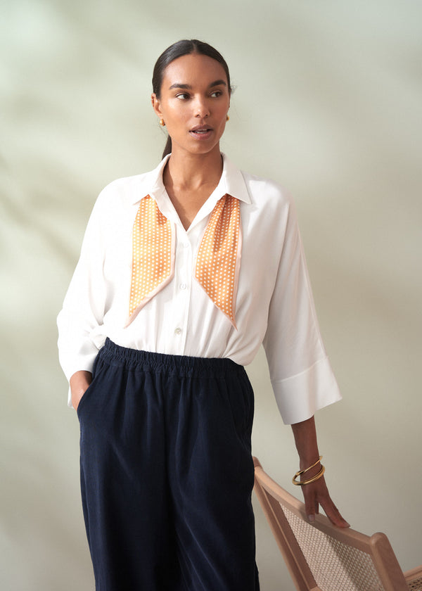 Woman wearing a white blouse with orange accents and dark pants against a plain background