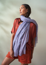 Woman wearing a red shirt and orange shorts with a blue and white striped scarf on a plain background