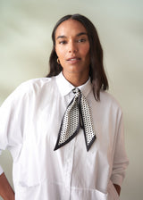 Woman wearing a white shirt with a black and white polka dot scarf around her neck against a plain background