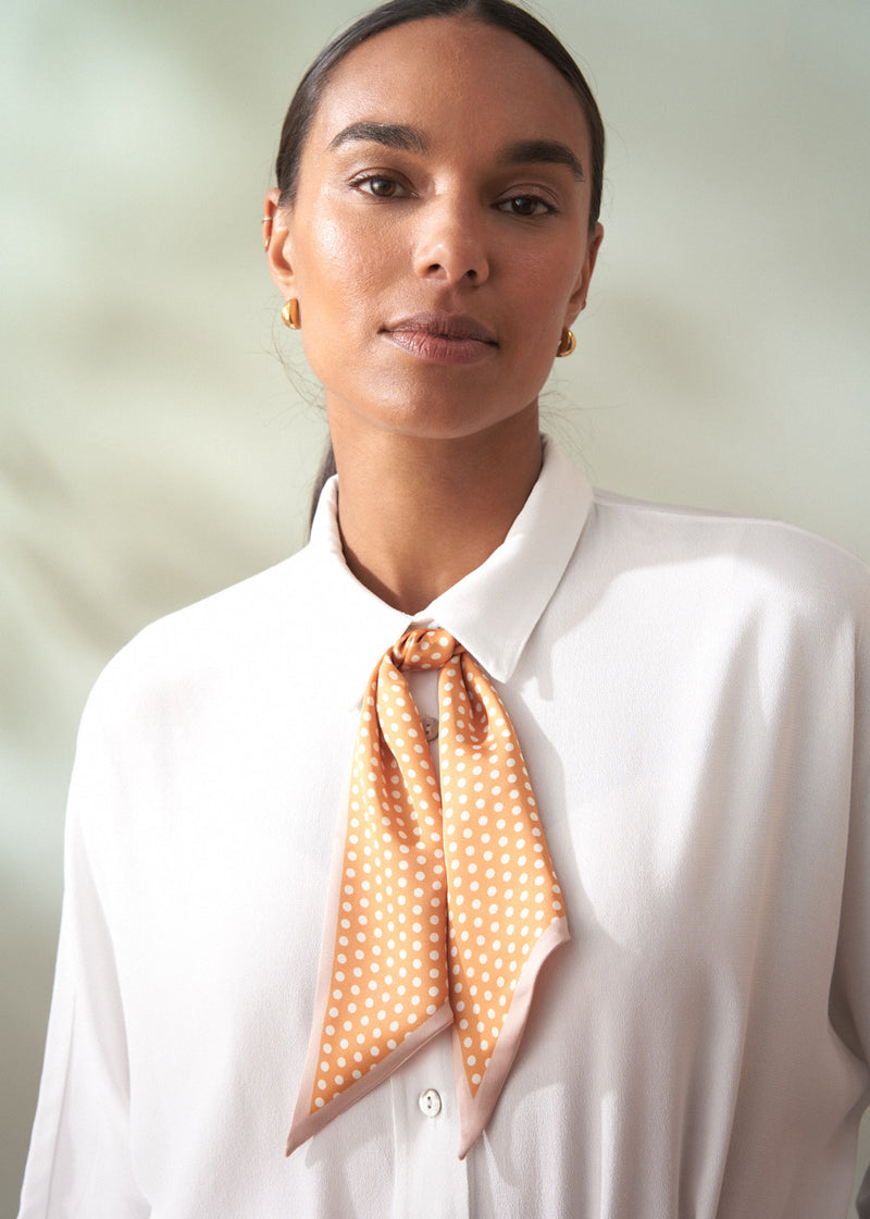 Woman wearing a white shirt with an orange polka dot scarf around her neck against a light background
