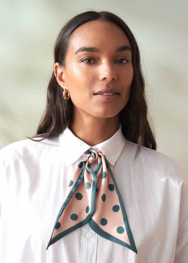 Woman wearing a white shirt with a green and pink polka dot scarf around her neck against a light background