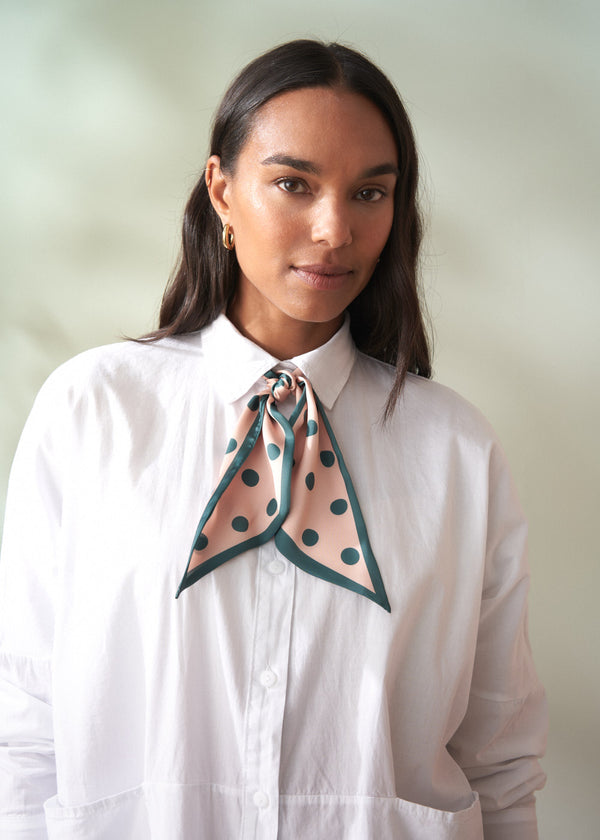 Woman wearing a white shirt with a green polka dot scarf around her neck against a plain background