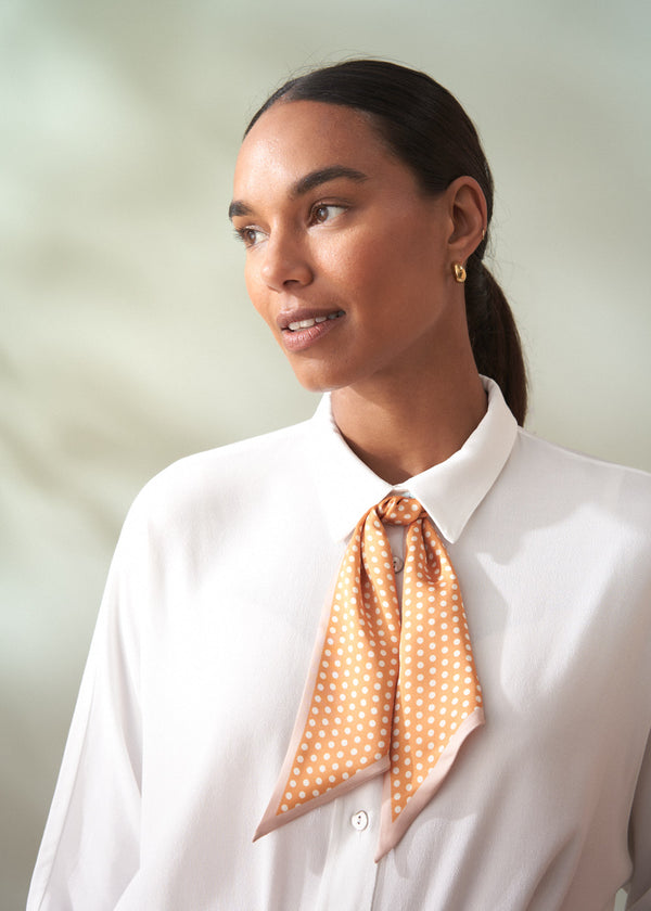 Woman wearing a white shirt with an orange polka dot scarf around her neck against a light green background