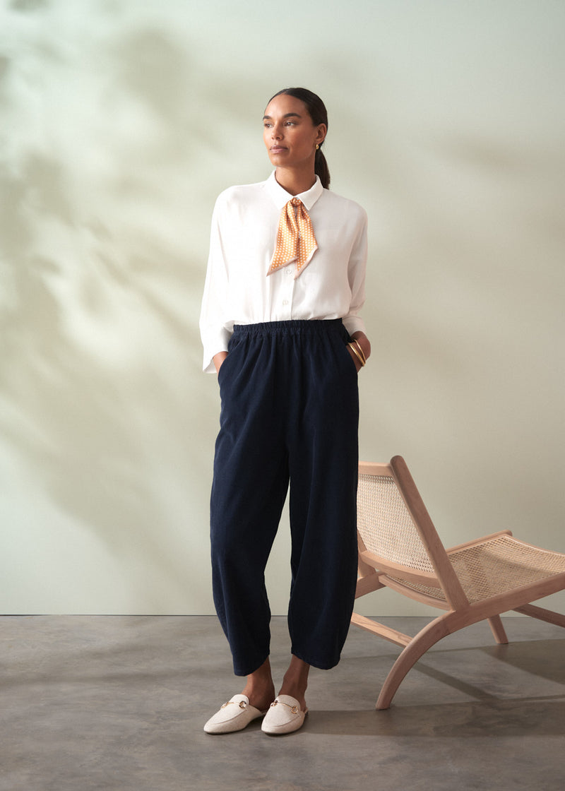 Woman wearing a white blouse with a yellow tie and navy pants standing in a room with a chair.